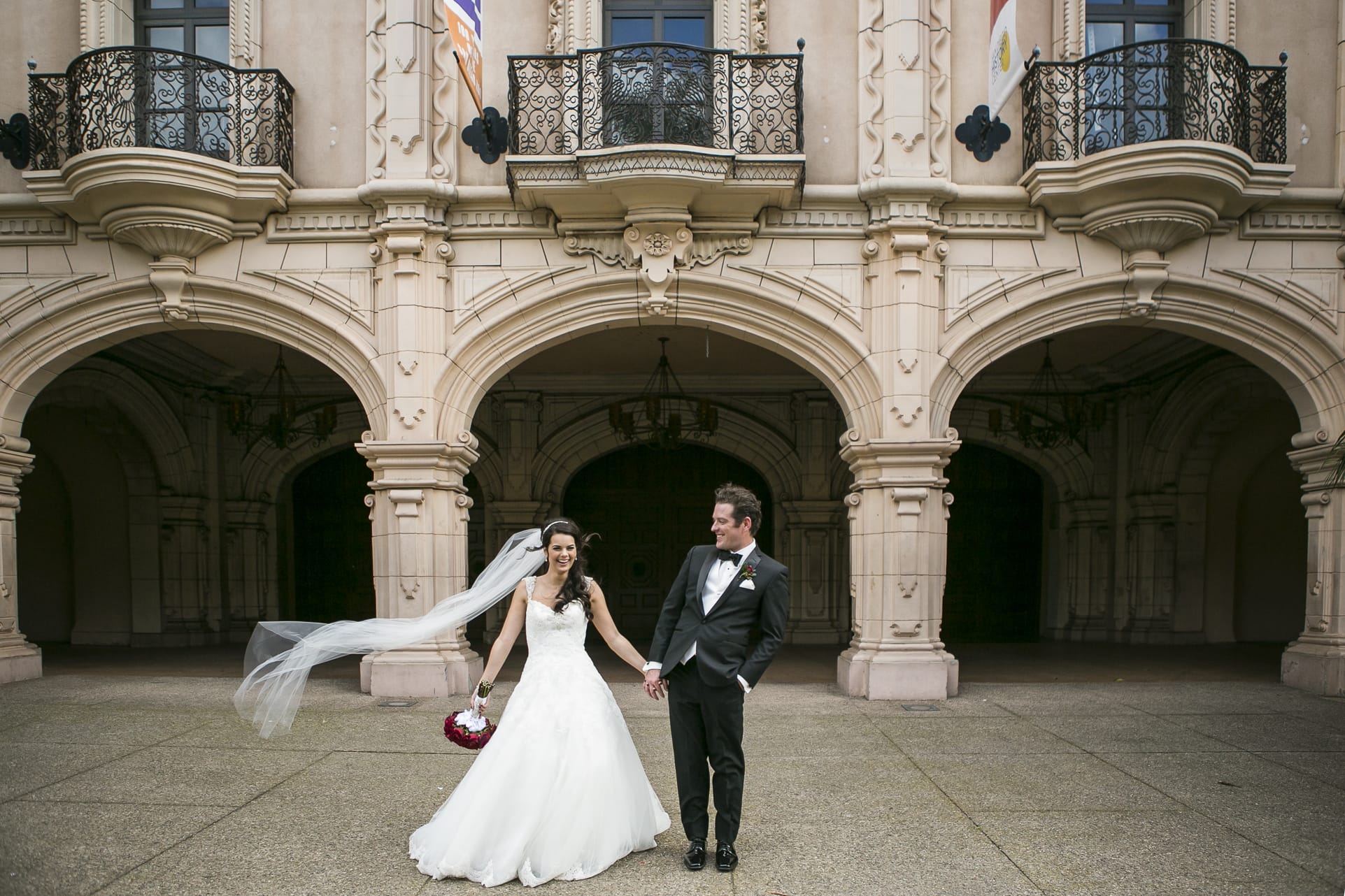A bride and groom stand hand in hand outside a grand building with ornate arches and balconies. The bride wears a flowing white dress and veil that flutters in the wind, while the groom is dressed in a black suit. Both are smiling and looking to their left, captured beautifully by their San Diego wedding photographer.