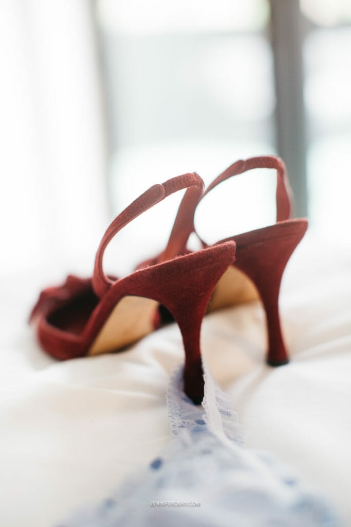 Close-up photo of a pair of red high-heeled shoes with open backs, placed on a bed with soft lighting. A piece of light blue lace fabric is draped in front of the shoes. The background is out of focus.