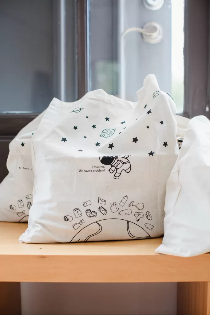 A white tote bag sits on a wooden surface with a minimalist design featuring space-themed illustrations, including stars, planets, and an astronaut. The text reads, "Houston, We have a problem." Captured by a San Diego Branding Photographer, the background shows a corner of a gray door with a metallic handle.