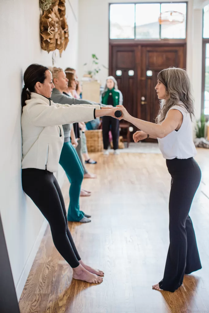 A group of people participate in an indoor fitness class. One instructor assists a participant who performs a wall exercise using a bar. The room, captured by a San Diego Branding Photographer, has wooden floors, large floor-to-ceiling windows, and a door in the background.