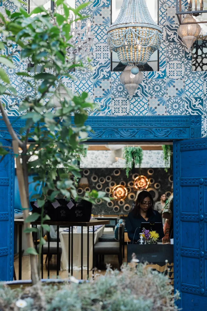 A woman sits at a table at Ettan restaurant with intricate blue and white patterned walls, reminiscent of a Palo Alto Bat Mitzvah. A large blue door frames the scene with a hanging chandelier in the center. Plants and green foliage add a touch of nature to the interior decor.