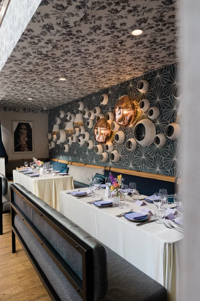 A sophisticated dining area set for a shabbat dinner at Ettan restaurant in palo Alto, California. Tables set with white tablecloths, neatly arranged utensils, and glassware. The walls, adorned with geometric patterns and circular mirrors, complement the floral arrangements. This elegant setting is perfect for hosting a Palo Alto Bat Mitzvah.