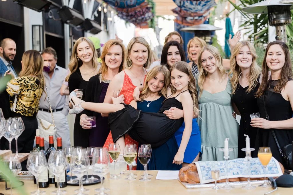 A group of smiling people posing at an outdoor gathering in Palo Alto. In the foreground, a girl in a black dress is being playfully held by an adult at what looks like a Bat Mitzvah celebration. The table in front of them holds various drinks, glasses, and decorative items. Vibrant decorations adorn the background.