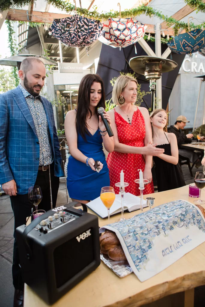 A group of people stands around a table set with wine glasses, challah, candles, and a challah. One person holds a microphone, and others look engaged in the moment. They are outdoors under a decorated canopy with greenery and lights at their shabbat dinner at Ettan restaurant in palo Alto, California