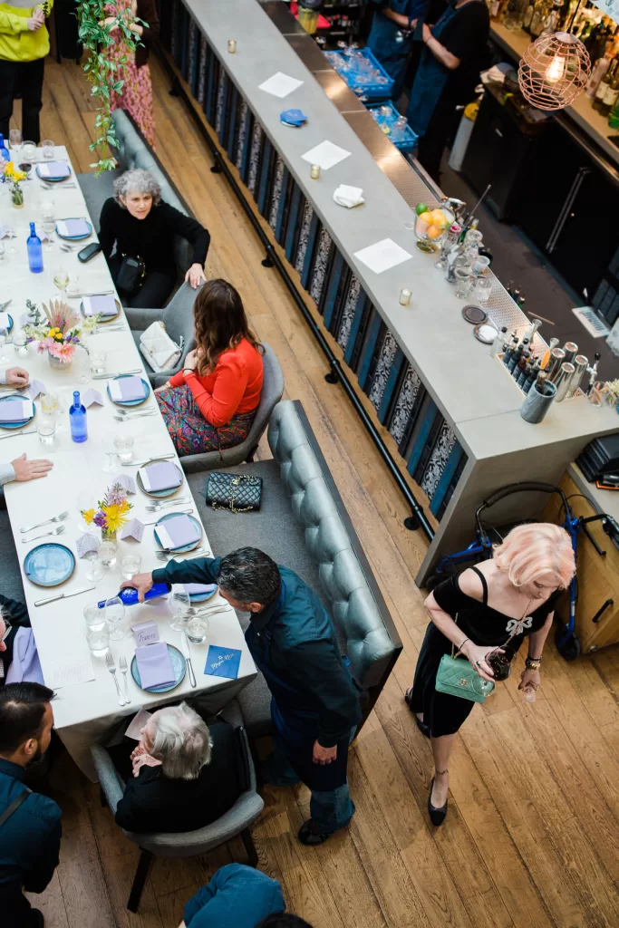 A bustling restaurant shabbat dinner at Ettan restaurant in palo Alto, California features a long table set for eight people, celebrating a Bat Mitzvah. Four diners are seated and conversing while others mill around, including fames event planner marcy blum walking by. The table is adorned with flowers and set with plates, glasses, and menus.
