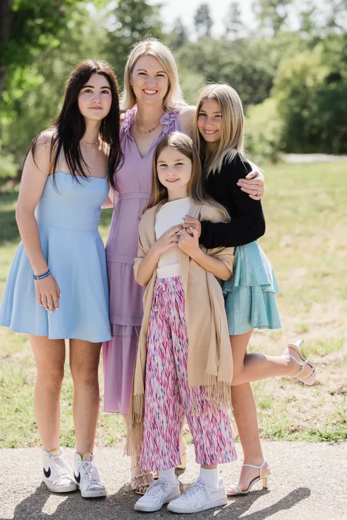 A group photo of four females standing outdoors on a sunny day captures a joyful Palo Alto Bat Mitzvah moment. An adult woman in a lilac dress stands in the center, with three girls of varying ages around her. The girl on the left wears a blue dress, the girl on the right a green dress, and the youngest in front wears a patterned skirt with a shawl. Trees and