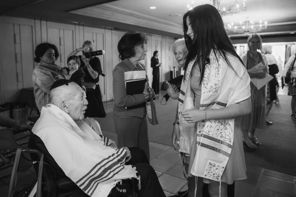 A black and white photo shows an elderly man seated in a wheelchair, wearing a prayer shawl and kippah. He faces a young girl also wearing a prayer shawl, likely at her Palo Alto Bat Mitzvah. Several people stand around them, engaged in conversation, with some holding books or taking photographs.