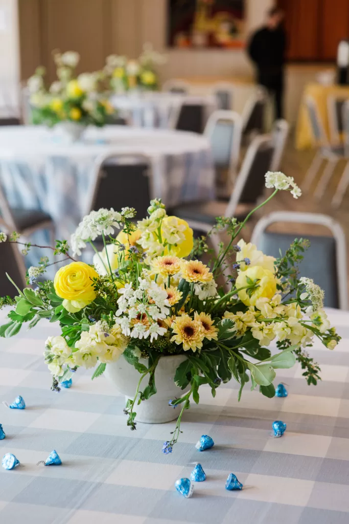 A table centerpiece of fresh yellow and white flowers in a white vase is placed on a table with a blue and white checkered tablecloth. Blue wrapped chocolates are scattered around. In the background, there are additional floral arrangements and empty chairs, ready for the Palo Alto Bat Mitzvah celebration.