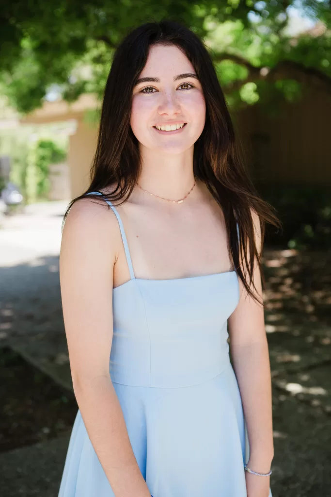 A young woman with long dark hair smiles at the camera during a Palo Alto Bat Mitzvah. She is wearing a light blue sleeveless dress and a simple necklace. The background is an outdoor setting with greenery and a pathway.
