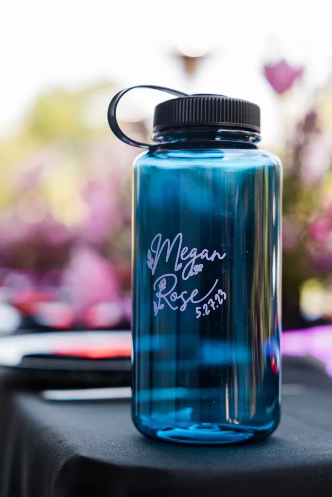 A blue water bottle with a black cap sits on a table. The bottle has white text that reads "Megan & Rose 5.27.23" with floral designs, commemorating their Palo Alto Bat Mitzvah. The background shows slightly blurred pink and green foliage.
