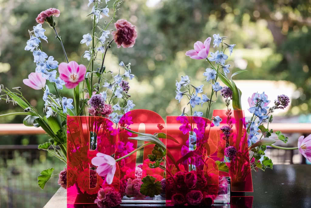 A colorful floral arrangement featuring pink tulips, blue delphiniums, and assorted greenery is artistically presented behind translucent red letters spelling "BABY." Set against a lush, green outdoor background, it creates a vibrant and cheerful atmosphere perfect for any Palo Alto Bat Mitzvah.