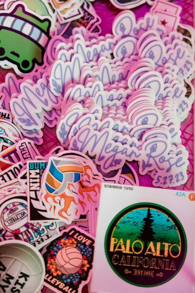 A colorful collection of stickers is scattered on a pink surface. Prominent ones include custom stickers with "Megan Rose" and a date, celebrating her Palo Alto Bat Mitzvah, along with a volleyball image and a "Palo Alto California" design. Other random stickers display various images and text.