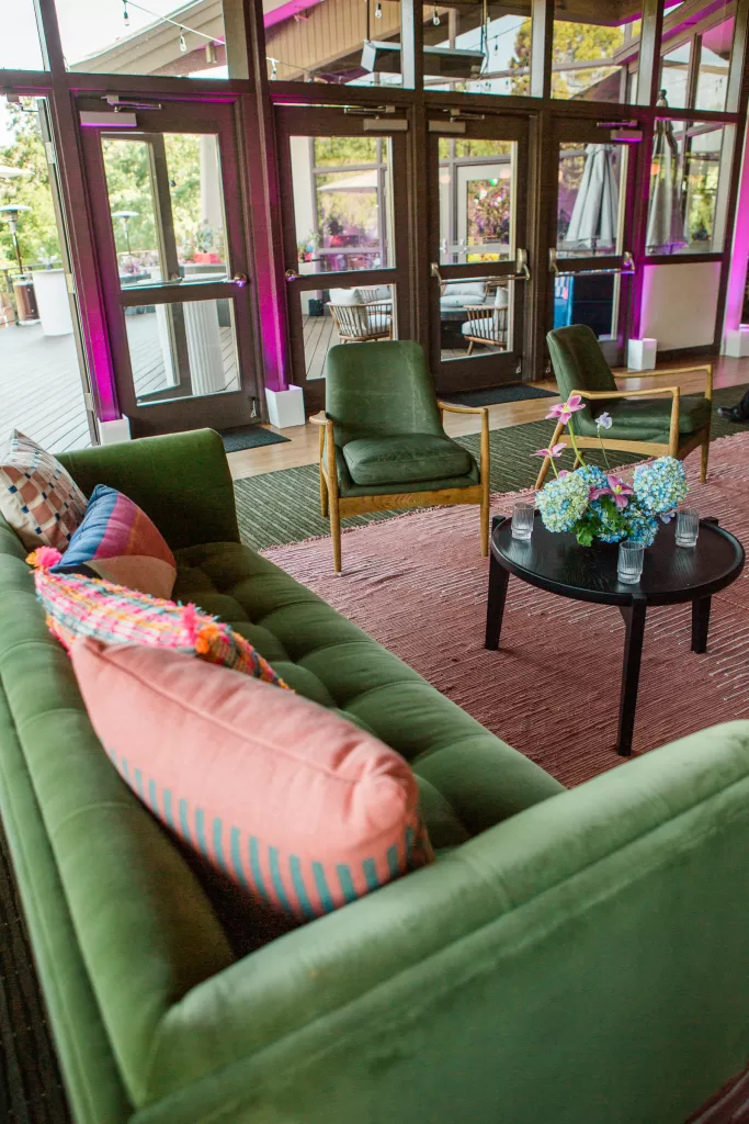 A stylish, modern indoor seating area perfect for a Palo Alto Bat Mitzvah, featuring a green velvet couch with colorful cushions, two matching green chairs, a black round coffee table with fresh flowers, and glass doors in the background with vibrant pink lighting outside.