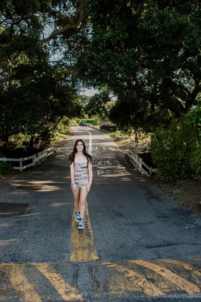A young woman with long hair walks down a tree-lined road in Palo Alto. She wears a colorful strapless top and shorts with sneakers. The road is shaded by foliage, and a yellow line runs down the center. The background includes a white wooden fence and overhanging branches, creating an enchanting setting reminiscent of her Bat Mitzvah day.