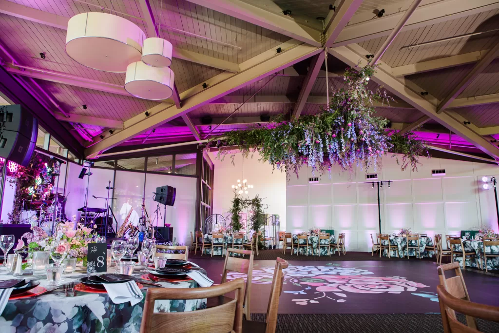 A beautifully decorated banquet hall hosts the Palo Alto Bat Mitzvah, with floral arrangements hanging from the ceiling, round tables adorned with elegant place settings, and soft purple lighting. The dance floor features a floral design, and a stage with musical equipment is set up.