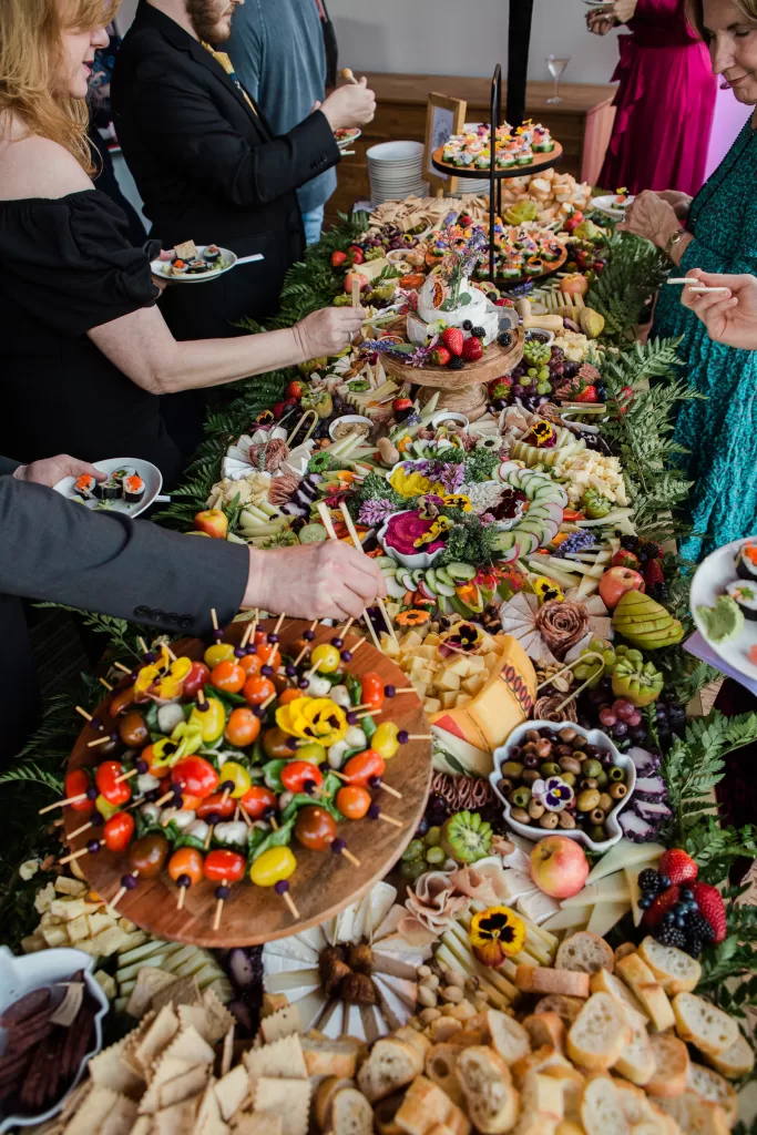 A lavish buffet spread at a Palo Alto Bat Mitzvah features an array of colorful appetizers, including skewered vegetables, assorted cheeses, fruits, olives, sliced bread, and crackers. Several people are serving themselves from the table adorned with decorative greens and floral accents.