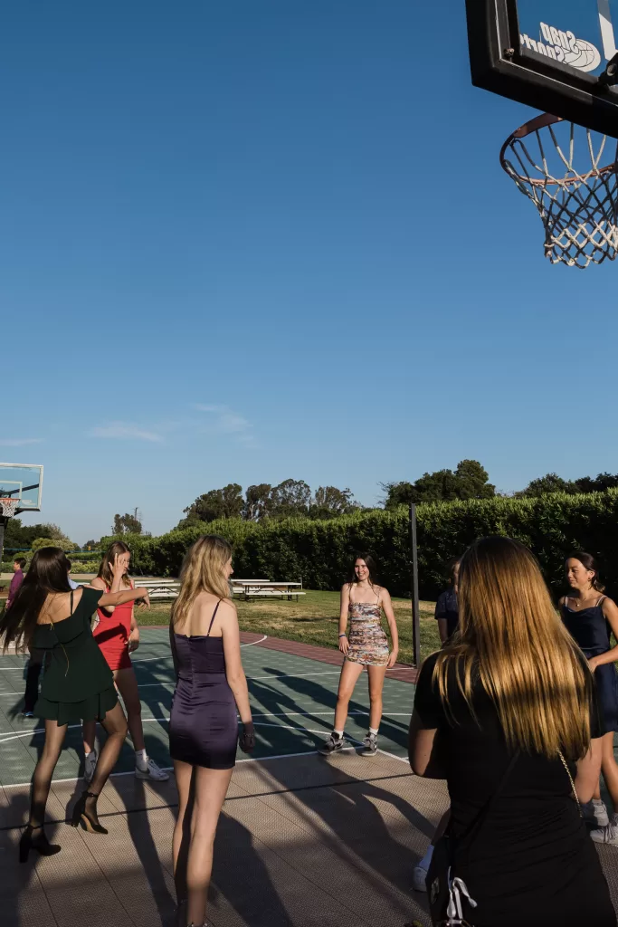 A group of women is playing basketball on an outdoor court under a clear blue sky, perhaps gathering after a Palo Alto Bat Mitzvah. Some are wearing casual dresses and others in sportier attire. They appear to be enjoying themselves, with trees and greenery visible in the background.