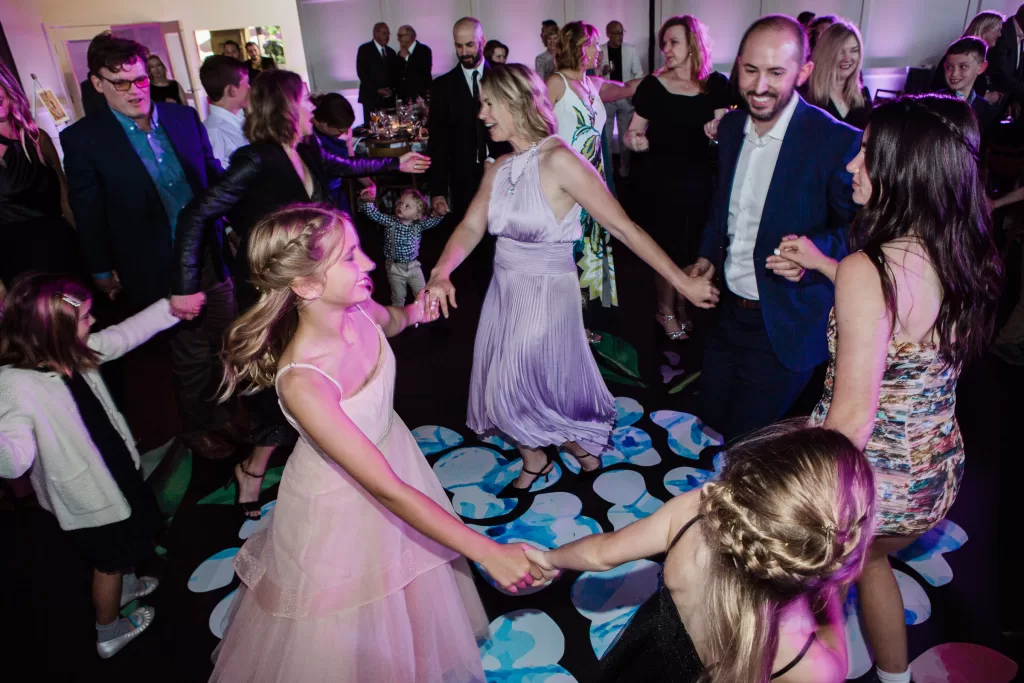 A lively group of people, including children and adults, dance together in a colorful and joyous setting at a Palo Alto Bat Mitzvah. They hold hands, forming a circle on an illuminated dance floor. Everyone is dressed in formal attire, and the ambiance suggests a festive celebration.