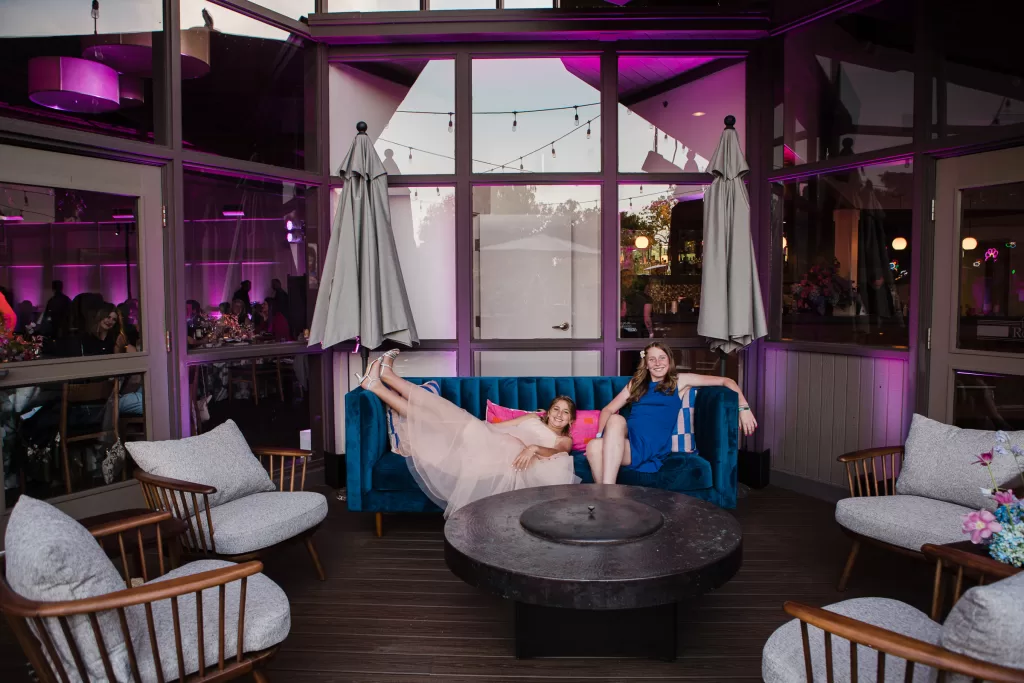 Two people are lounging on a blue couch in a modern, open-air seating area with purple lighting, reminiscent of a chic Palo Alto Bat Mitzvah. One person is lying down with legs playfully kicked up, while the other is sitting upright. Surrounding the sofa are patio chairs and tables, with a round firepit in the center.