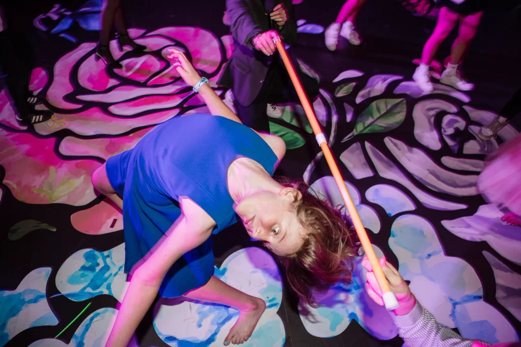 A person in a blue dress bends backward in a limbo dance under a colorful light at a Palo Alto Bat Mitzvah. They attempt to pass under a horizontal stick held by two people. The floor is decorated with a vibrant, abstract floral pattern.