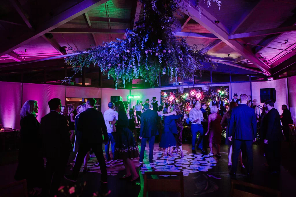 People are enjoying a dance floor under dim, colorful lighting at a Palo Alto Bat Mitzvah. A large floral arrangement hangs from the ceiling, and the room is decorated with vibrant, gradient lights that create a festive atmosphere. Guests are mingling and dancing in pairs and groups.
