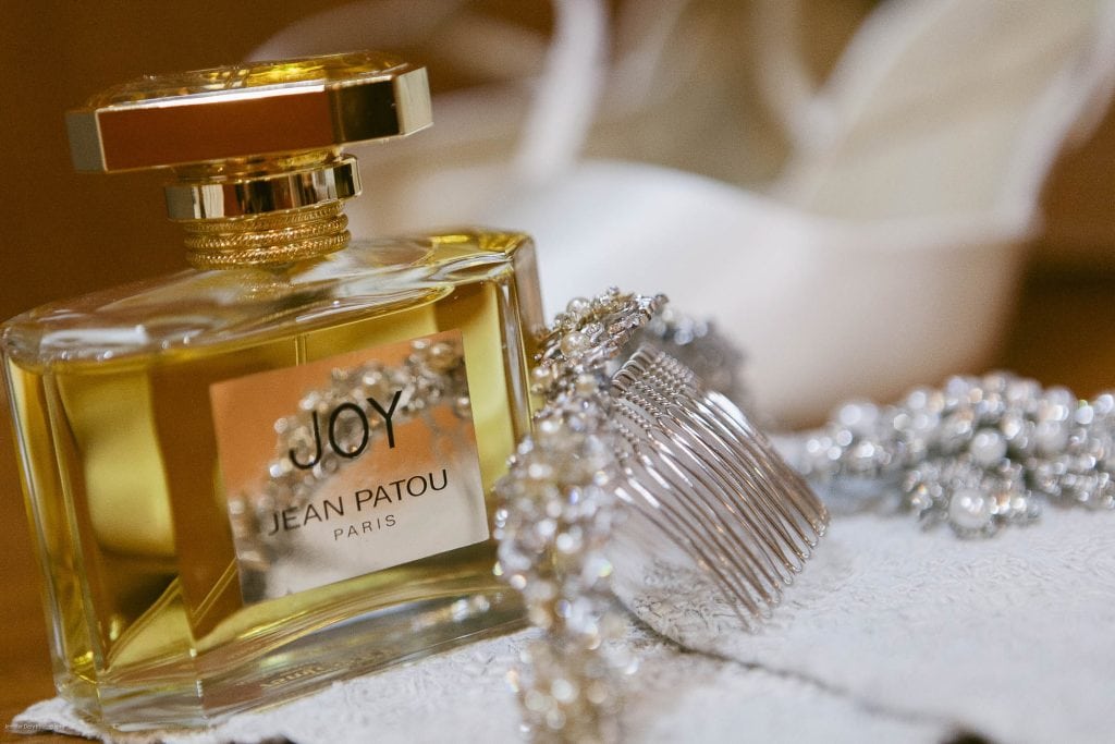 A bottle of JOY by Jean Patou perfume sits on a white surface next to a sparkling jeweled hair comb and silver jewelry, with a blurred white shoe in the background.