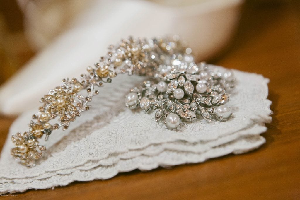 A sparkling silver and pearl brooch and a jeweled hair accessory with floral designs rest on a delicate, white lace fabric atop a wooden surface.