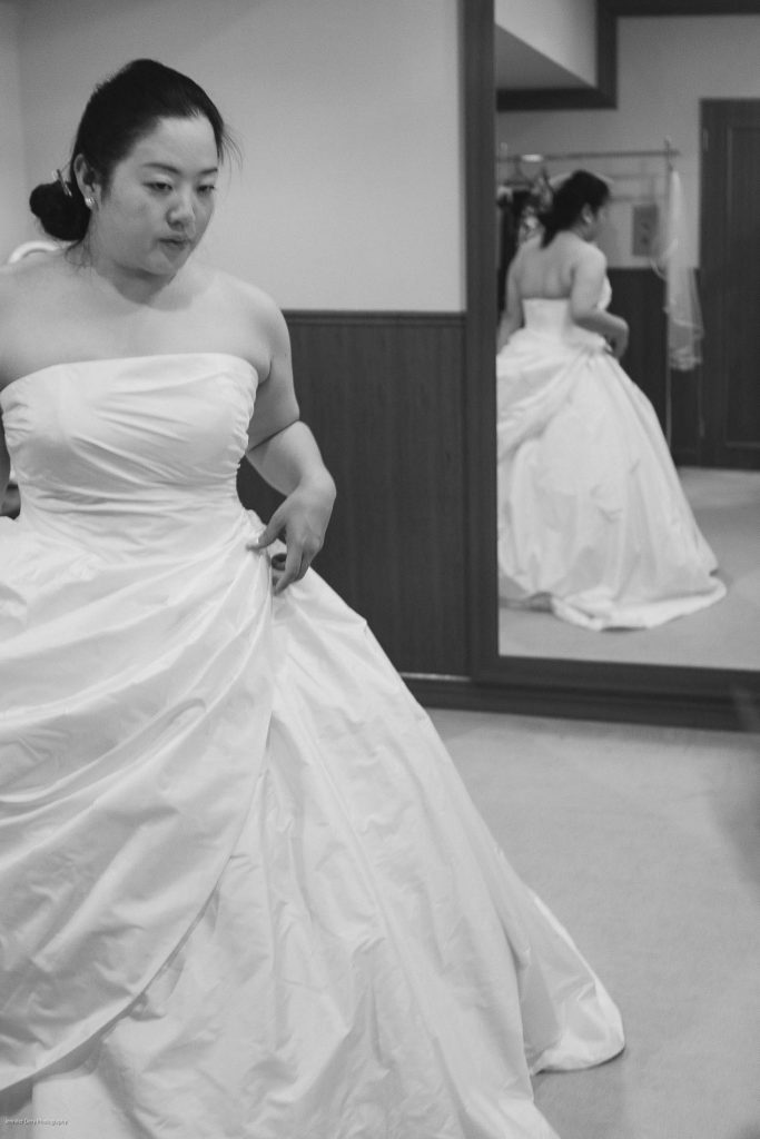 A woman in a strapless white wedding dress stands in a room, adjusting the skirt. Her reflection appears in a large wall mirror behind her. The photo is in black and white.