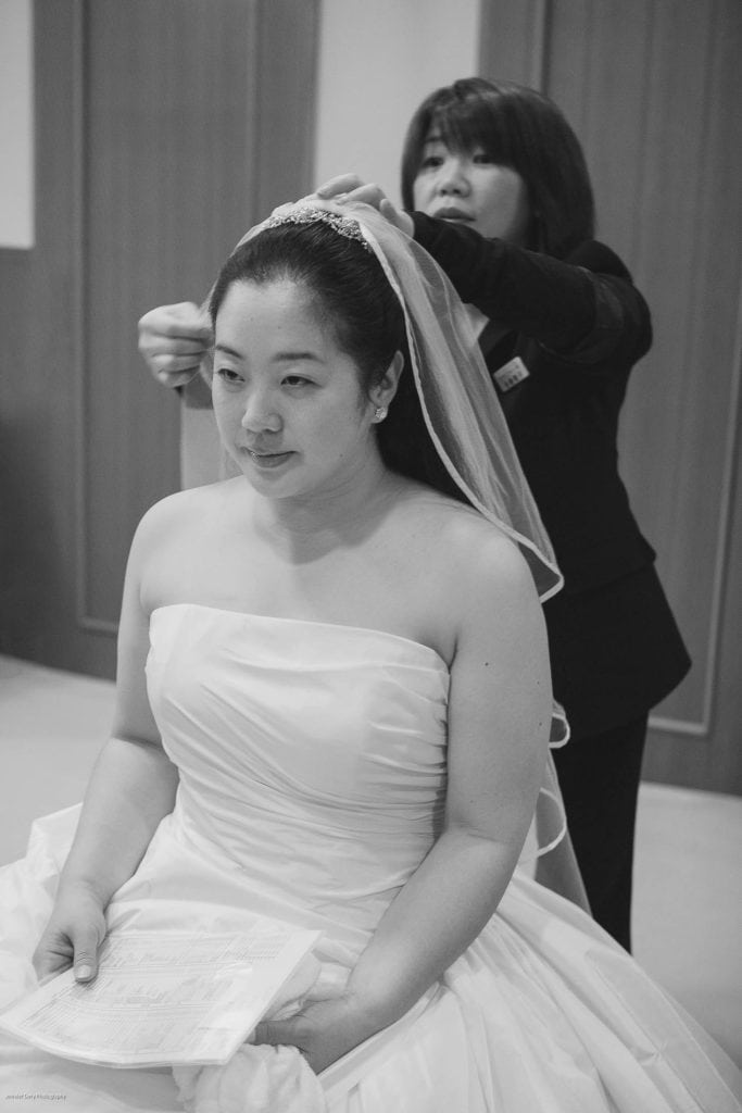 A bride in a strapless wedding gown sits holding papers while another woman adjusts her veil from behind. The scene appears to be indoors, with both women focused on their tasks. The image is in black and white.