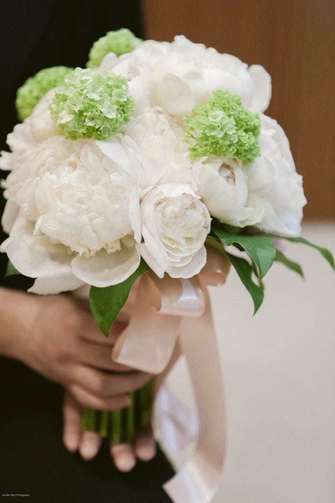 A person holding a bouquet of white peonies and green hydrangeas, tied with a pale pink ribbon. The background is softly blurred.