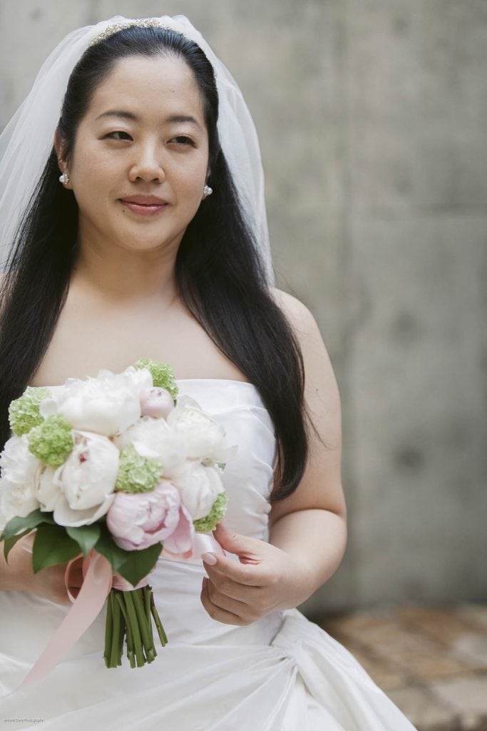 A bride in a white dress holds a bouquet of white and pale pink flowers with greenery and a pink ribbon. She has long dark hair, a veil, and pearl earrings, and stands in front of a neutral, blurred background.