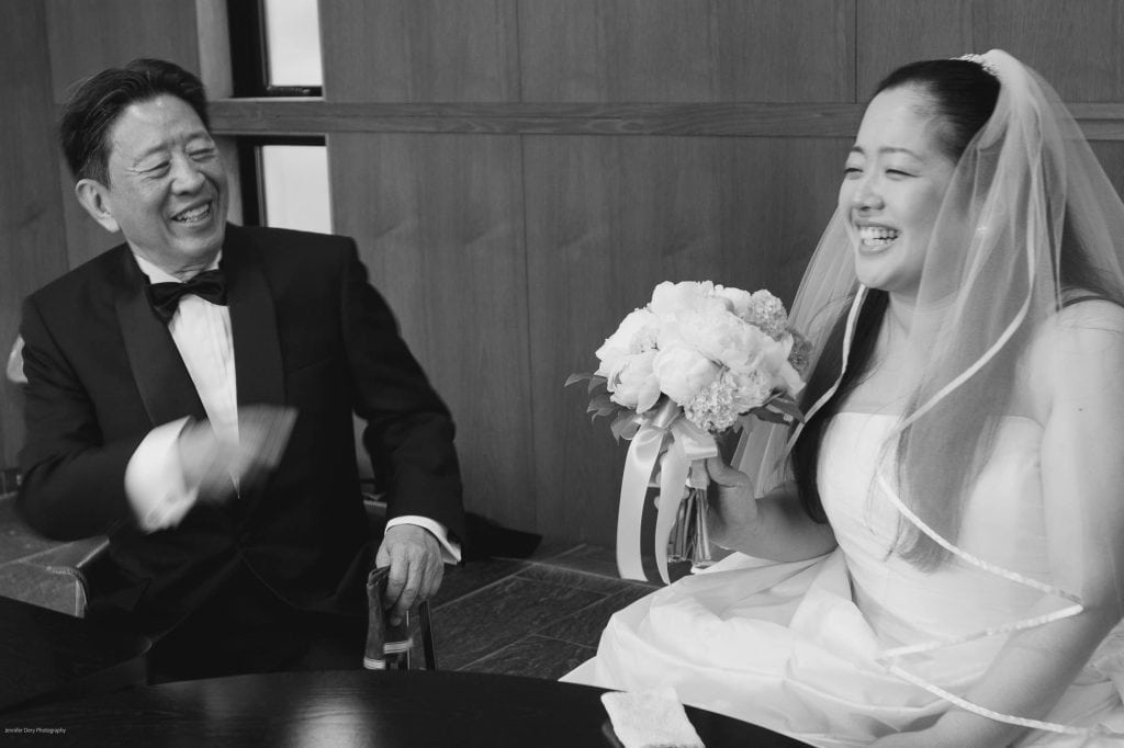 A bride in a white dress and veil holding a bouquet smiles while sitting next to an older man in a tuxedo. Both are laughing and appear happy, sharing a joyful moment together indoors.