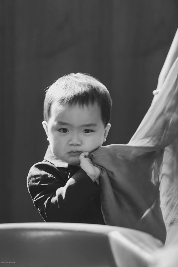 A young child with short hair, wearing a dark outfit, looks directly at the camera while holding onto a draped piece of fabric. The image is in black and white with soft lighting and a blurred background.