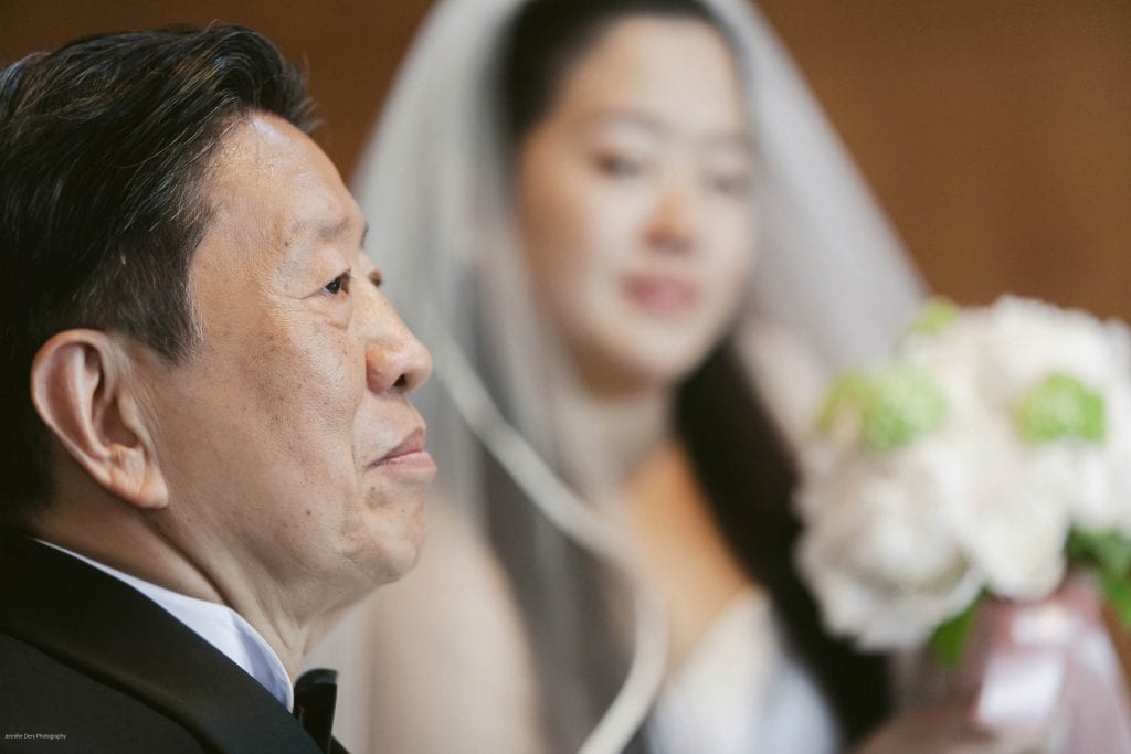 A close-up of an older man in a suit looking thoughtful, with a bride in a white dress and veil holding a bouquet softly blurred in the background.