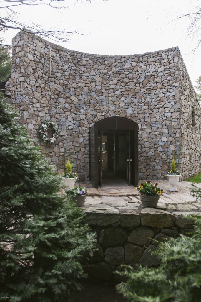 A curved stone building with an arched double doorway, decorated with a flower wreath and two potted plants. A stone wall and green bushes are in the foreground.