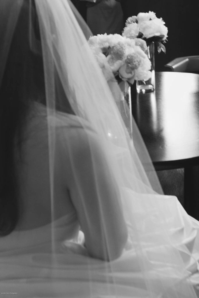 A black and white photo of a bride from behind, wearing a veil and strapless gown, sitting near a table with vases of flowers. The scene is softly lit and elegant.