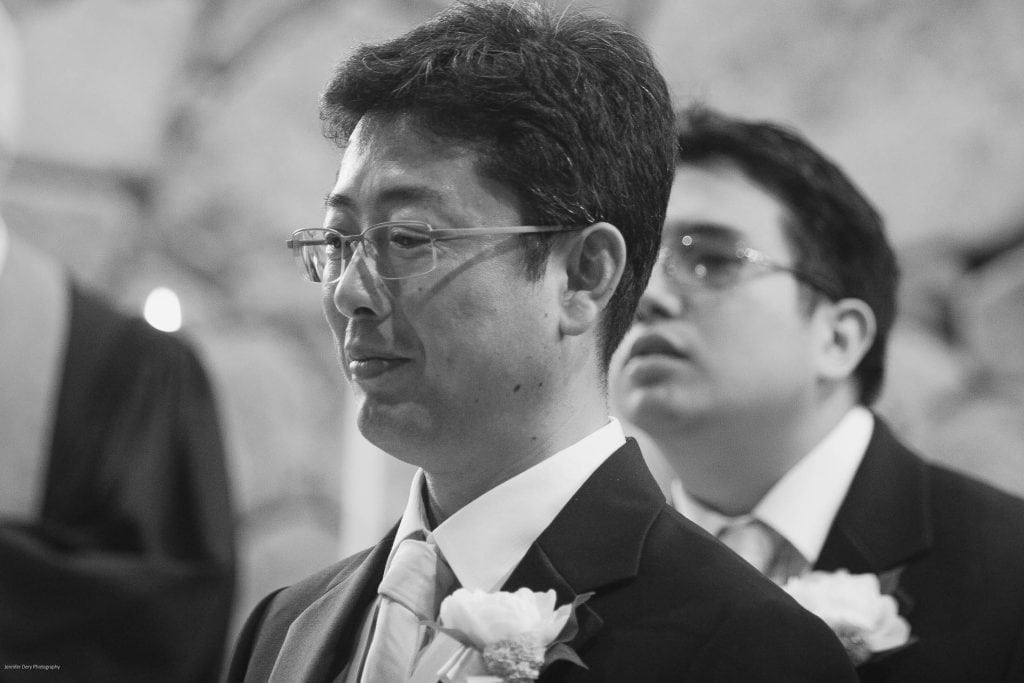 Two men in suits, each wearing glasses and a boutonniere, stand side by side at what appears to be a formal event, possibly a wedding. The image is in black and white.