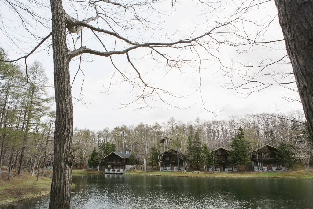Several wooden cabins line the far side of a calm lake, surrounded by tall trees with sparse branches, under a cloudy sky in a serene, natural setting.