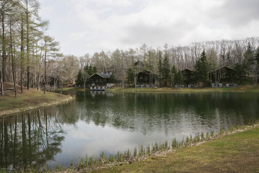 A calm pond reflects trees and wooden cabins on its far shore, with bare branches and evergreens under a cloudy sky in a peaceful, natural setting.