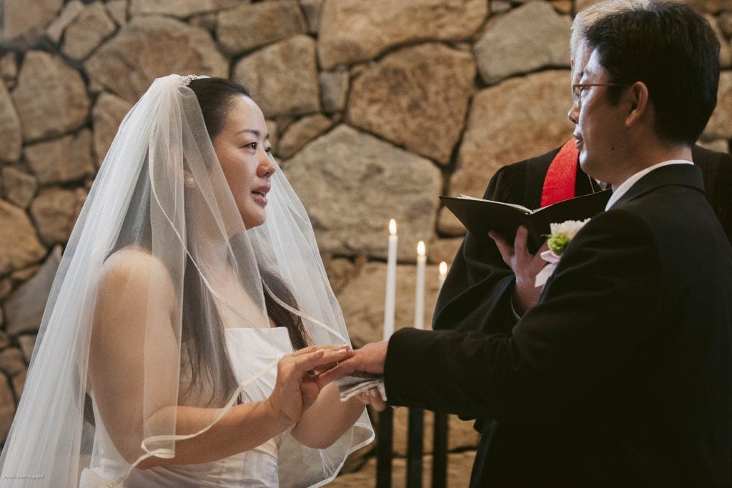A bride in a white dress and veil places a ring on the groom’s finger during a wedding ceremony, with a stone wall and lit candles in the background.