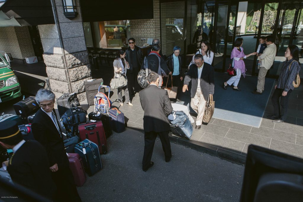 A group of people stands outside a building entrance with several suitcases and bags on the ground. Some individuals are picking up luggage, while others are conversing or waiting. A hotel staff member assists the guests.