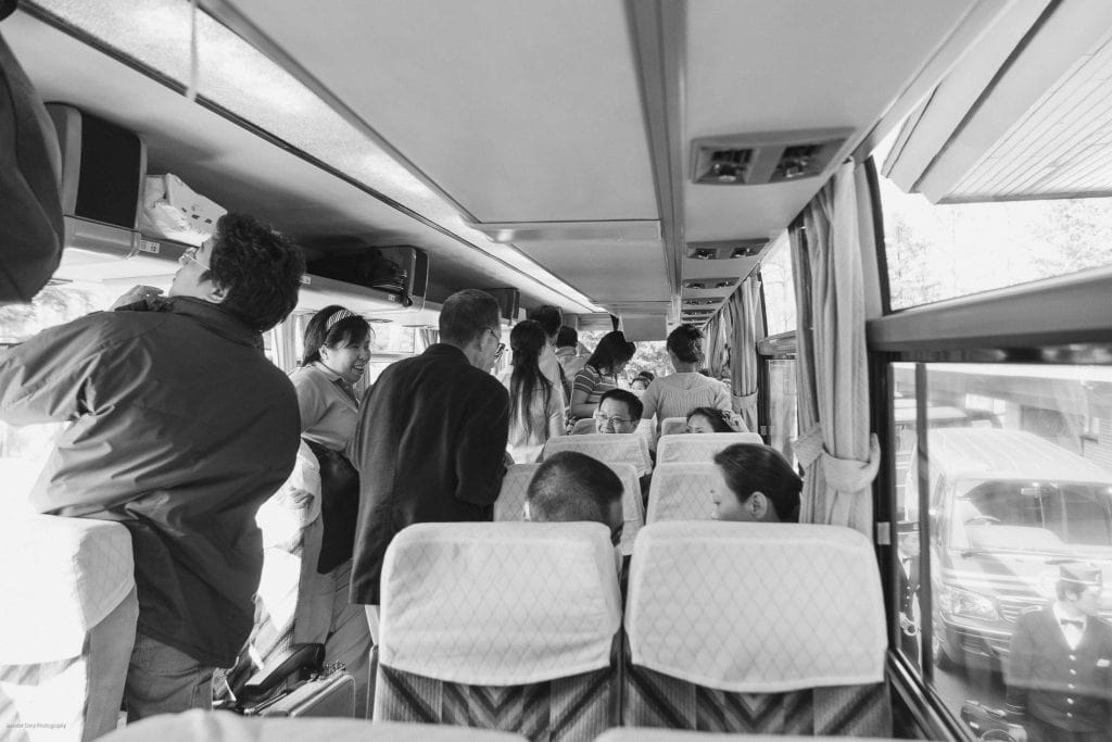 A black and white photo showing people standing and sitting inside a bus, some placing bags in overhead compartments, while others talk or look out the windows. The scene captures a busy, crowded atmosphere.