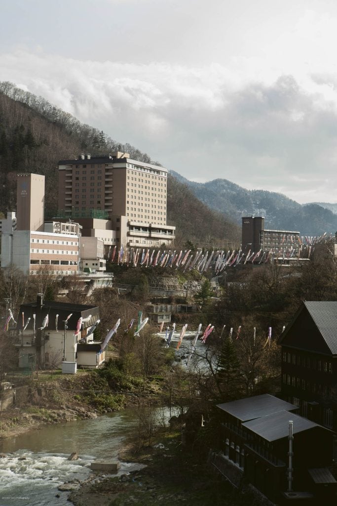 A riverside town with tall buildings, colorful carp streamers flying across the river, and forested mountains in the background under a partly cloudy sky.