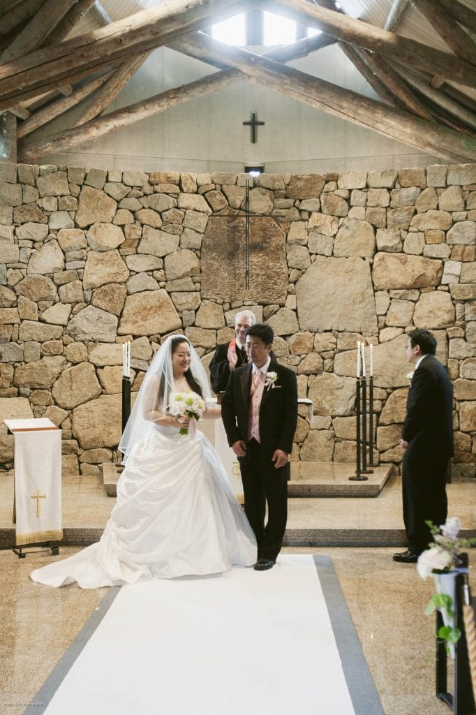 A newlywed couple walks down the aisle of a rustic stone chapel, smiling. The bride wears a white gown and veil, holding a bouquet, while the groom is in a black suit. Two men stand nearby, one clapping.