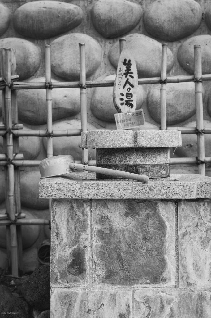 A metal ladle rests on a stone basin in front of a bamboo fence and a wall of large, rounded stones. A wooden sign with Japanese writing stands upright behind the basin. The image is in black and white.