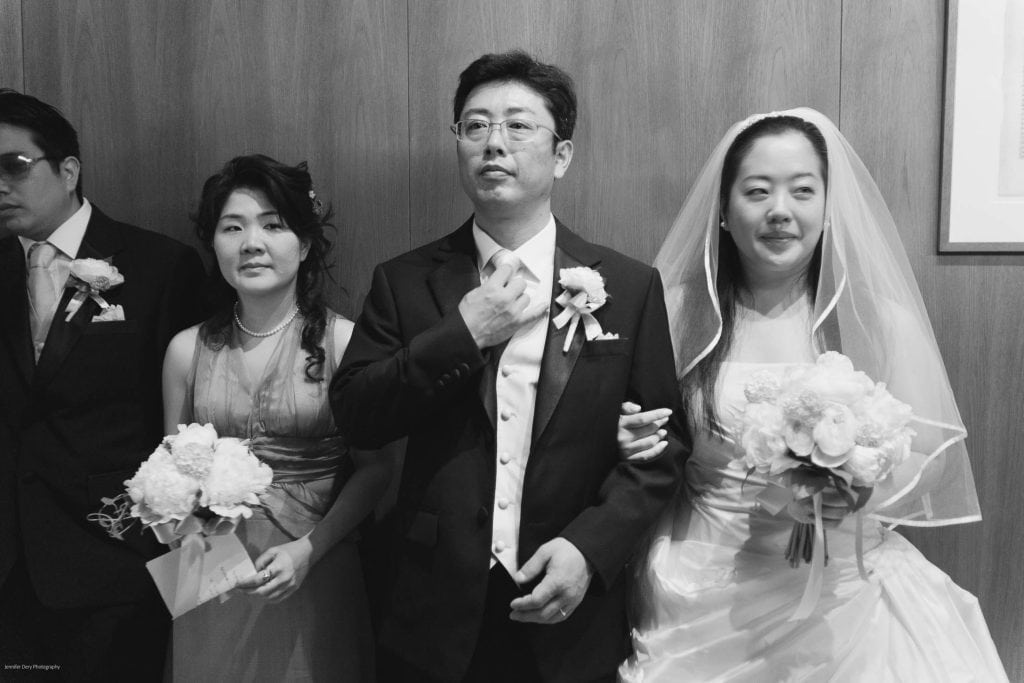 A black and white photo of four people dressed formally, including a bride holding a bouquet, a groom adjusting his tie, and two attendants standing beside them, all in front of a wooden wall.