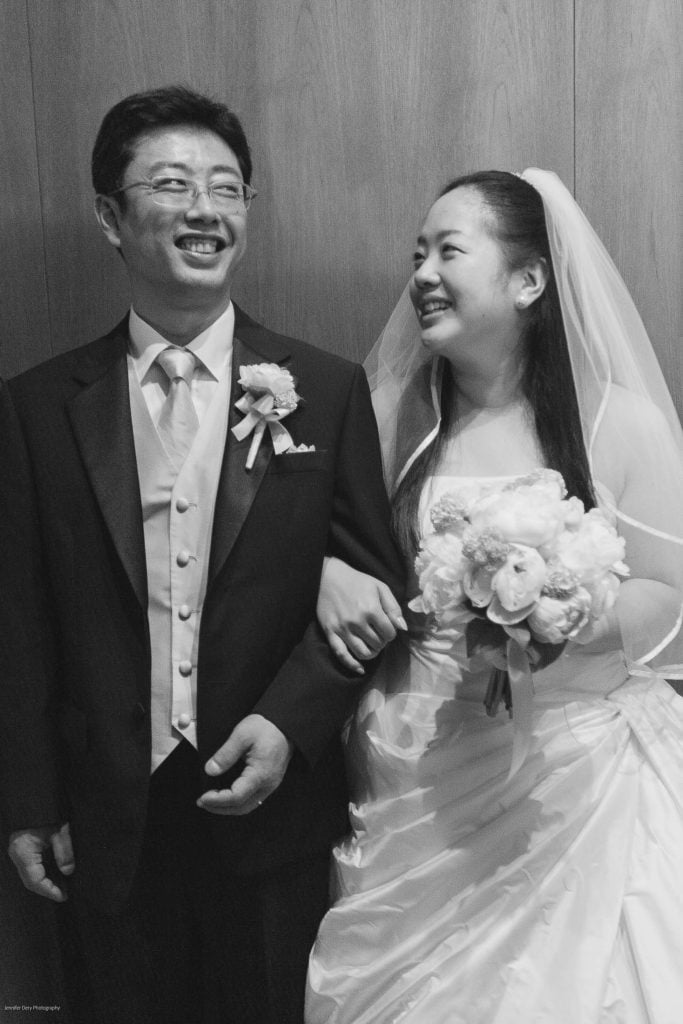 A smiling bride and groom stand arm in arm. The bride, wearing a veil and holding a bouquet, looks at the groom, who is in a suit with a boutonniere. Both appear joyful against a plain background.
