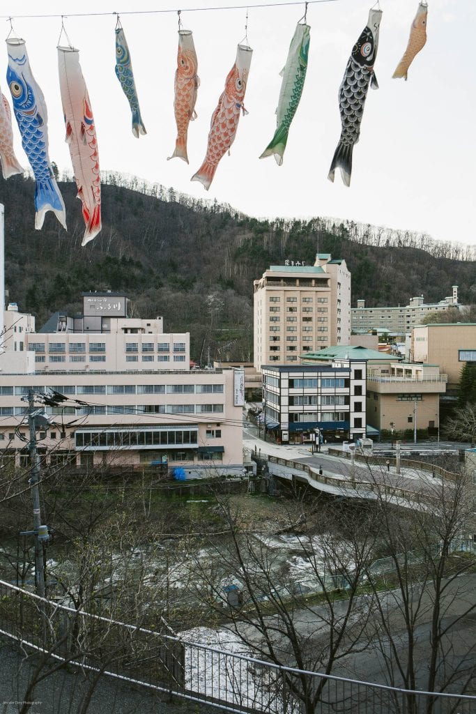 Colorful koi-shaped windsocks (koinobori) hang above a small cityscape with modern buildings, a river, bare trees, and forested hills in the background under a cloudy sky.