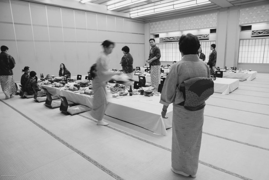A black-and-white photo of people in traditional Japanese attire arranging food on low tables in a tatami-mat room. Some individuals are seated, while others are serving or organizing the meal.