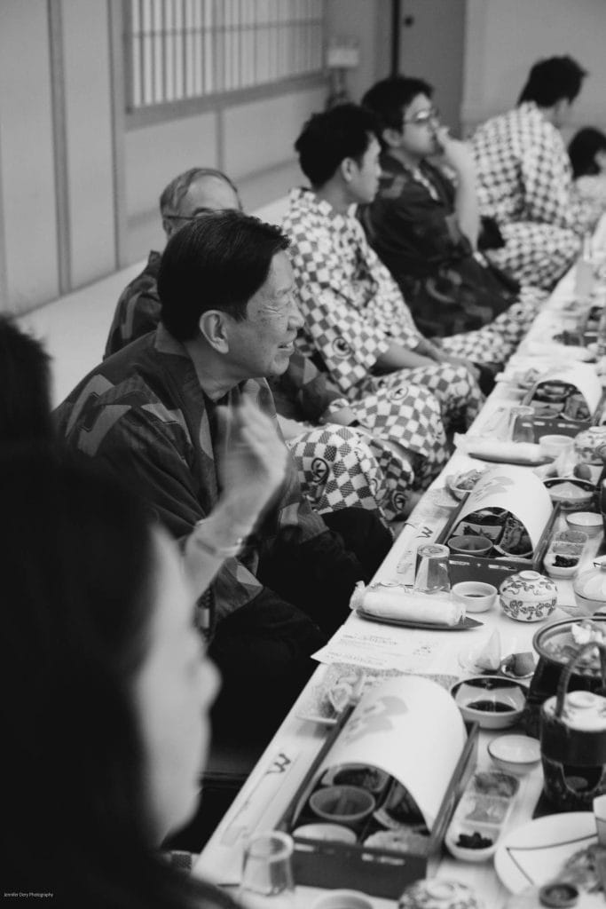 A group of people wearing traditional Japanese robes sit closely together on the floor around a long, low table set with various dishes and bowls, engaged in conversation. The photo is in black and white.